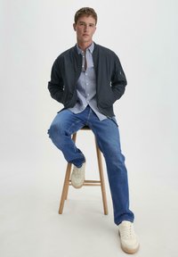 Schwarze Bomberjacke, blau gestreiftes Hemd mit Knöpfen, blaue Jeans und beigefarbene Sneaker. Das Modell sitzt auf einem Holzstuhl vor einem schlichten Hintergrund.