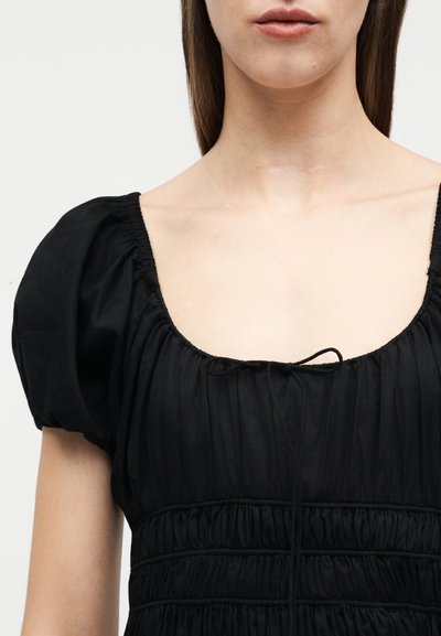 Γυναίκα που φοράει μαύρο φόρεμα με κοντά μανίκια, με ρυτιδωμένο μπούστο και μικρό φιόγκο στο τετράγωνο ντεκολτέ, εμφανιζόμενη από τους ώμους μέχρι τα χείλη.