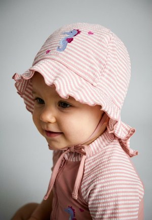 Toddler in pink striped sun hat with seahorse embroidery and matching outfit, looking slightly down and smiling softly.