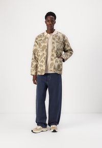 Camo-mønstret jakke i beige og grøn, parret med en lys beige sweater, løse mørkeblå jeans og beige sneakers med grå accenter.