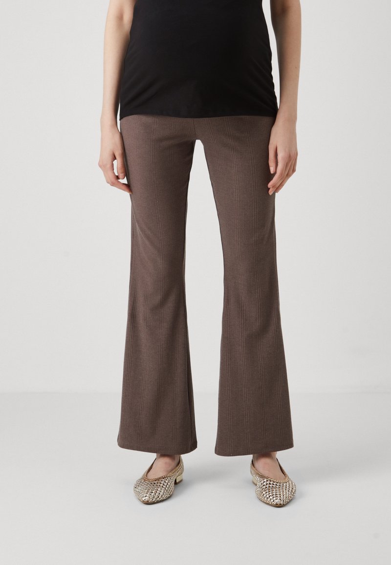 Kvinna i bruna ribbade flare-byxor, svart topp och guldiga flätade ballerinas står på vit golv och bakgrund.