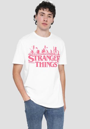 Junger Mann in einem weißen T-Shirt mit pinkem "Stranger Things"-Logo und Silhouetten von Kindern, die über dem Text Fahrrad fahren, kombiniert mit blauen Jeans.