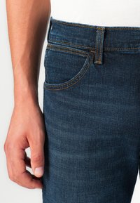 Jeans de denim azul escuro com costura visível e um bolso frontal. O tecido tem uma leve textura, e a cintura apresenta um pequeno detalhe em forma de loop.