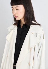 Manteau trench crème avec un col en cascade, boutons double boutonnage, et une chemise noire élégante en dessous. Texture lisse et coupe ajustée.