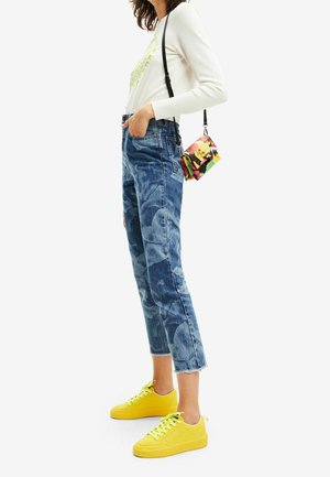 Kobieta ubraną w biały top z długim rękawem, niebieskie jeansy z wysokim stanem w kwiatowy wzór, jasne żółte trampki oraz z małą, kolorową kwiatową torebką na ramię.