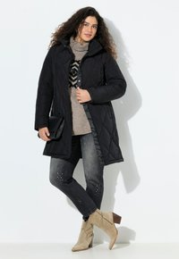 Kvinna i svart vadderad kappa, beige mönstrad tröja, mörkgrå jeans med dekorationer, beige mocka ankelstövlar och håller en svart kuvertväska.
