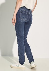 Dark blue skinny jeans with a mid-rise waist, back pockets, and a textured denim finish, paired with white and green sneakers.