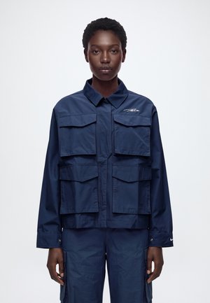 Un mannequin se tient devant un fond blanc, vêtu d’une veste utilitaire bleu marine à quatre poches à rabat et d’un pantalon assorti.