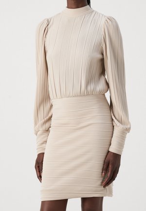 Femme portant une robe en maille côtelée beige avec de longues manches bouffantes et une jupe cintrée sur un fond blanc uni.