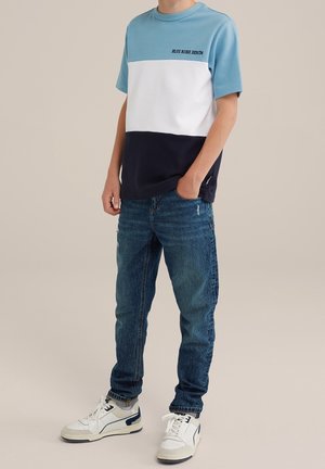 Persona che indossa una maglietta a blocchi di colore blu, bianco e navy con la scritta "Blue Ridge Denim", jeans blu e sneakers bianche, in piedi contro uno sfondo neutro.