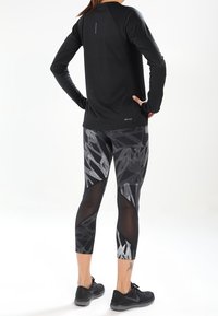 Zwart sporttop met lange mouwen en raglanmouwen; aansluitende legging met een patroon van zwart en grijs bladeren; zwarte sneakers.