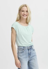 Mintgrünes Kurzarmoberteil mit rundem Halsausschnitt und subtilen Falten, kombiniert mit hellblauen, hoch taillierten Jeans.