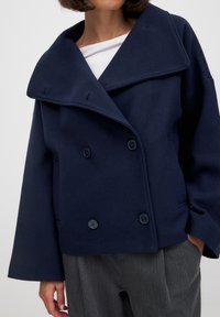 Tumesinine topeltkinnitatud villane mantel laia kraega, suurte nööpidega ja ülelahedaste varrukatega, millel on tekstuurne kangas ja esikotid.