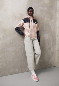 Mujer con un pañuelo negro, chaqueta en tonos pastel rosa y negra, pantalones beige y zapatillas rosas, de pie contra una pared de concreto liso.