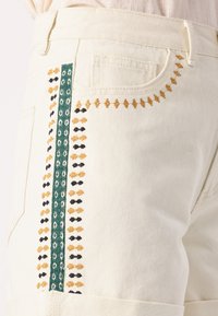 Gros plan sur un pantalon de couleur crème avec une rayure verticale verte brodée et de petites coutures décoratives orange et noires près de la poche.