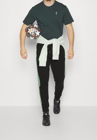 Mand iført sorte Adidas bukser og skjorte, som holder en sort-hvid Adidas fodbold under armen.