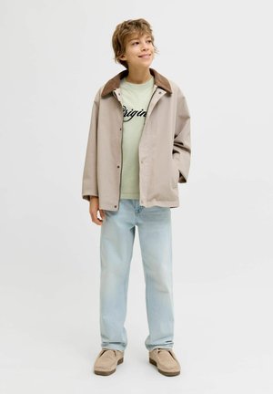 Usmívající se mladý člověk v béžové bundě s hnědým límcem, světle zelené košili s nápisem „Original“, světle modrých džínách a béžových botách stojí a dívá se doprava.