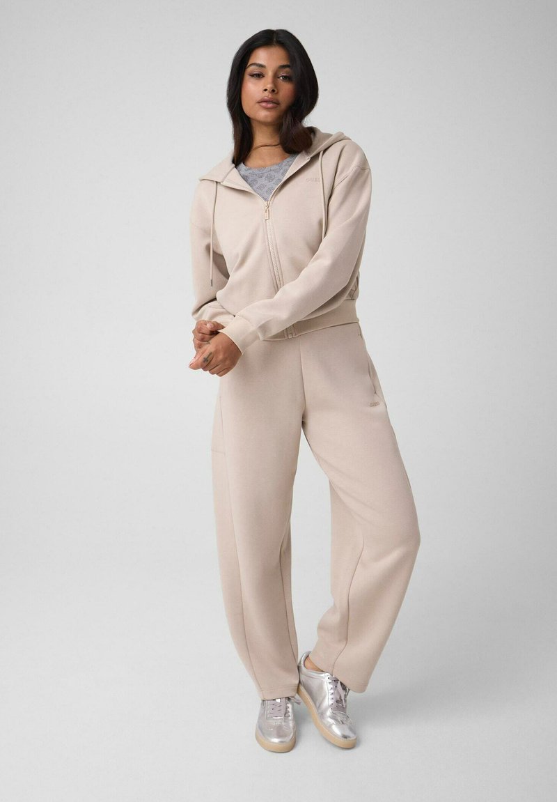Femme debout portant un sweat à capuche beige zippé, un pantalon large assorti, un t-shirt gris et des baskets argentées sur un fond gris uni.