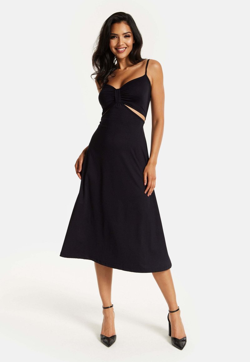 Black midi dress with thin straps, featuring a gathered bodice, cut-out sides, and a smooth, flowing fabric. High-heeled black shoes.