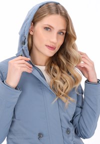 Kobieta z długimi blond włosami, ubrana w jasnoniebieską kurtkę z kapturem, poprawiająca kaptur, na białym tle.