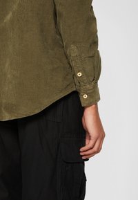 Bras et main droits détendus le long du corps, portant une chemise en velours côtelé vert olive avec poignet boutonné et un pantalon cargo noir avec rabat de poche.
