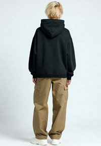 Felpa nera con un ampio cappuccio, abbinata a pantaloni cargo marrone chiaro con tasche laterali. La persona indossa l'outfit appoggiata a uno sfondo chiaro.
