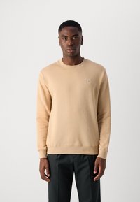Sudadera beige con cuello redondo y puños acanalados, que presenta un pequeño parche con el logo en la parte superior del lado izquierdo del pecho. Se lleva con pantalones oscuros.