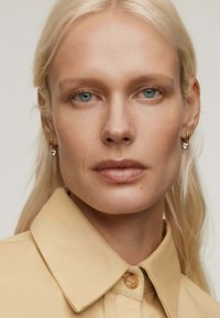 Camisa beige con cuello y cierre de botones, combinada con pendientes de aro dorados y un adorno de cristal colgante, todo con un fondo neutro.