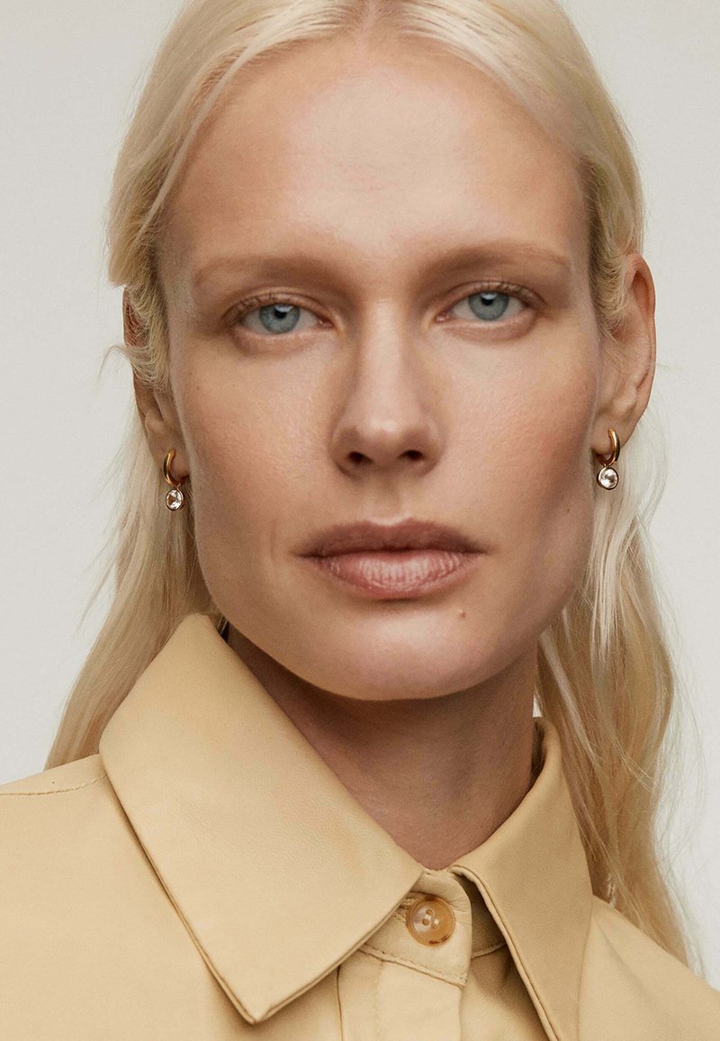 Camisa beige con cuello y cierre de botones, combinada con pendientes de aro dorados y un adorno de cristal colgante, todo con un fondo neutro.