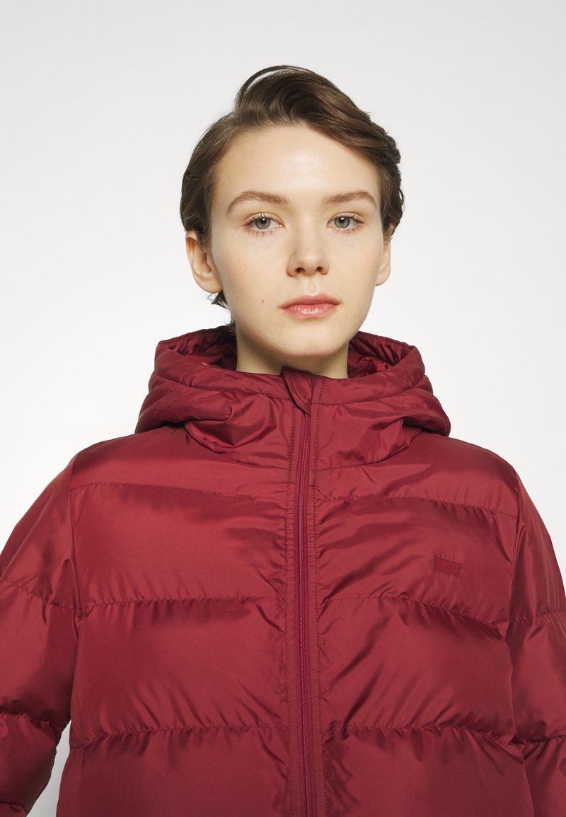 Giacca puffer bordeaux con cappuccio, caratterizzata da dettagli di cucitura e una zip frontale. Tessuto liscio e lucido con una texture trapuntata.