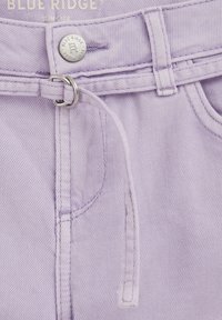 Nahaufnahme von lavendelfarbenen Jeans aus Denim mit Metallknopf, abgesteppten Taschen und einem dünnen Stoffgürtel, der durch doppelte Metallringe geführt ist.