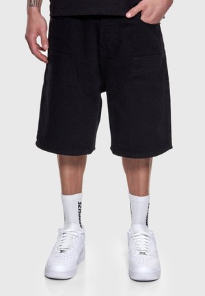 Mand iført sorte løse shorts, hvide crew-sokker med sort tekst og hvide sneakers, stående mod en enkel lys baggrund.