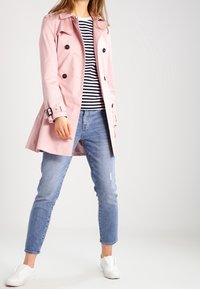 Rosa trenchcoat med svarta knappar, bälte i midjan och krage, bärs över en randig skjorta och blå slitna jeans, matchat med vita sneakers.