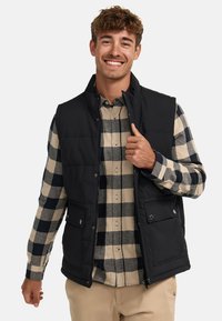 Chaleco negro con cuello alto sobre una camisa a cuadros, con bolsillos de presión y un diseño acolchado. El modelo está sonriendo y sosteniendo la cremallera.