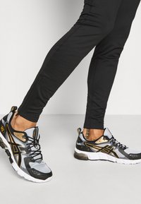 Schwarze und goldene Trainingsschuhe mit einem grauen Mesh-Obermaterial, ausgestattet mit einer strukturierten Sohle und auffälligen Logoakzenten an der Seite und Ferse.