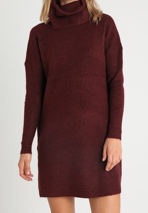 Abito a collo alto in maglia bordeaux con maniche lunghe e una vestibilità ampia. La texture è morbida e accogliente con un motivo a coste. Lunghezza fino a metà coscia.