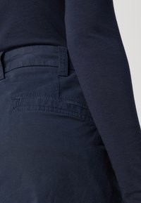 Person som bär mörkblå byxor med bakfickor och en matchande mörkblå långärmad topp, sedd från midjan till mitten av låret.