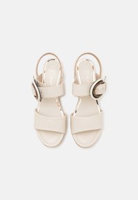 Beige sandaal met twee brede, overlappende banden en een slingback ontwerp. Bevat een ronde gesp als accent op de enkelband. Gladde textuur.