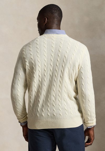 Mand iført en cremefarvet kabelstrikket sweater over en blå og hvidstribet skjorte med krave set bagfra mod en neutral baggrund.