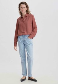 Rostfarbenshirt mit langen Ärmeln und Knopfleiste, kombiniert mit hellblauen, geraden Jeans und schwarzen Slingback-Ballerinas. Weicher Stofftextur.