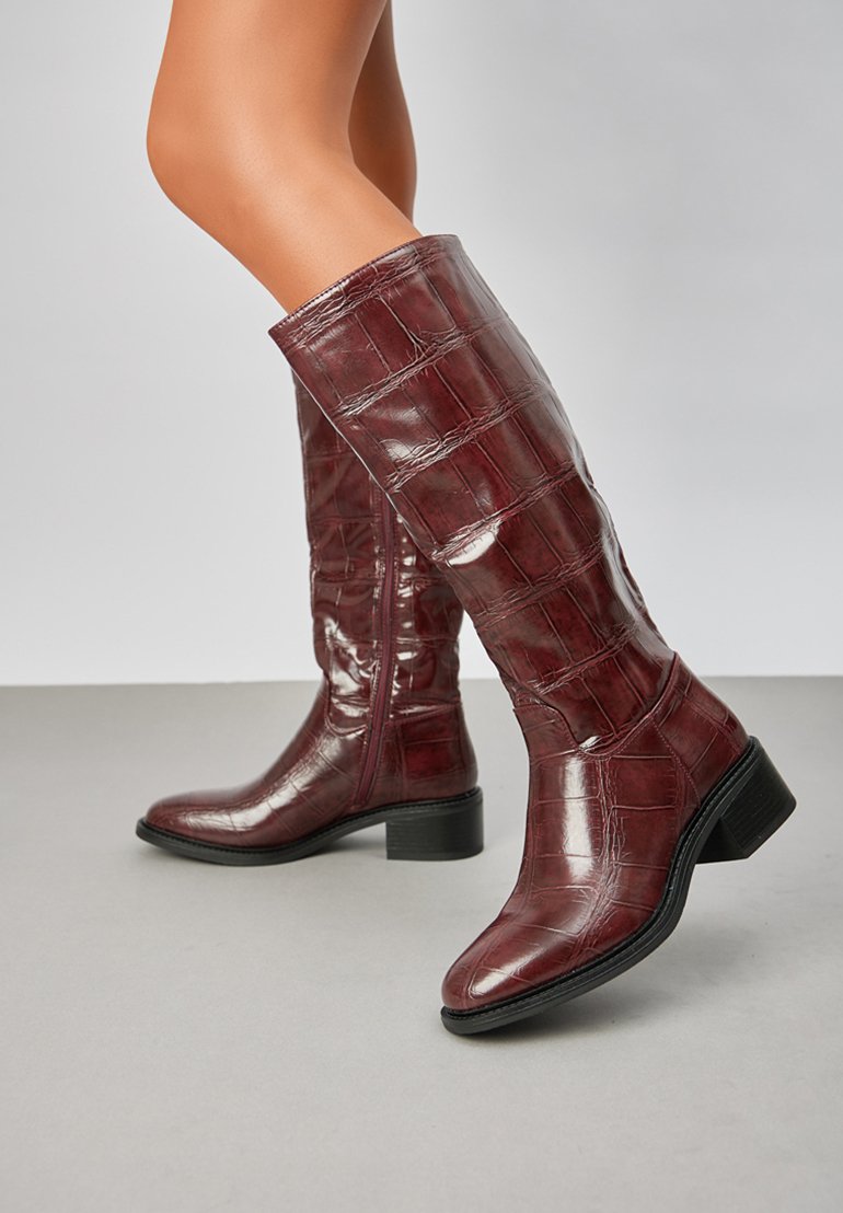Bottes bordeaux jusqu'au genou en matière brillante avec texture crocodile embossée, à bout carré et petit talon bloc, dotées d'une fermeture éclair sur le côté.