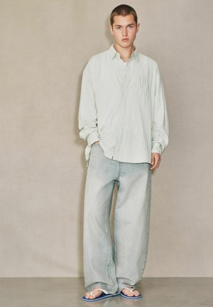 Young man standing against plain wall wearing oversized light shirt, wide-leg faded jeans, and blue flip-flops, hands in pockets, neutral expression.