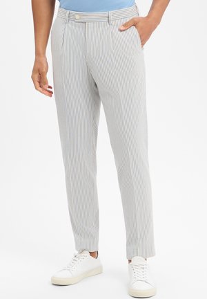 Person trägt hellblaue und weiße, vertikal gestreifte Hosen, kombiniert mit weißen Sneakers, steht mit einer Hand in der Tasche.