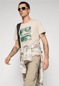 Mand iført beige Columbia T-shirt, solbriller, lysebrune cargo-bukser, fleece knyttet om livet, og en sort rygsækrem over skulderen.
