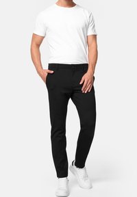 Pantaloni neri su misura con una texture liscia, dotati di tasche laterali, chiusura con bottone e vestibilità slim, abbinati a una maglietta bianca semplice e sneakers bianche.
