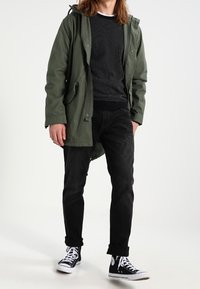Parka verde oliva con capucha sobre un suéter negro texturizado, combinado con jeans negros y zapatillas negras con cordones y suelas blancas.