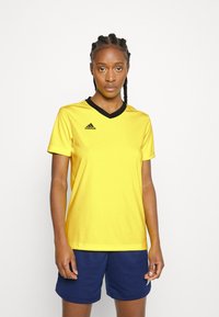 Geltonas sportinis marškinėliai su juodu V formos apykakle ir trumpomis rankovėmis, ant kairės krūtinės yra Adidas logo. Dėvimi su mėlynomis šortais.