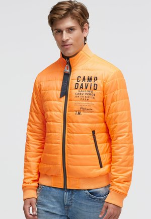 Mann trägt eine leuchtend orange gesteppte Jacke mit schwarzem Text und Reißverschlusstaschen, kombiniert mit blauen Jeans, steht vor einem einfarbigen Hintergrund.