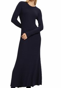Femme portant une longue robe en tricot côtelé bleu marine foncé, manches longues et corsage cintré, posant avec un bras plié derrière le dos.