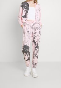 Tuta rosa con grandi disegni di personaggi in bianco e nero, composta da una giacca corta con zip e pantaloni elasticizzati. Il materiale appare morbido e casual.